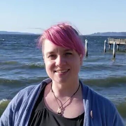 Person with pink hair smiling by a lakeside, wearing a blue jacket, sunny weather, wooden pier in the background.