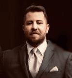 Man in a suit with a pink tie and pocket square against a dark background.